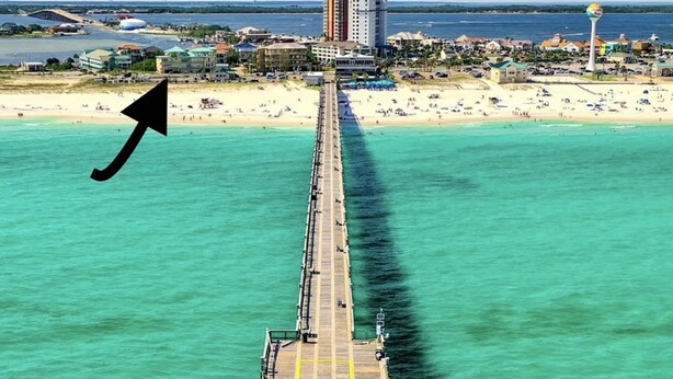 Une jetée s’avançant dans des eaux turquoise, avec une plage et des bâtiments en arrière-plan.