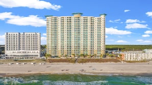 A beachfront hotel with multiple floors, a sandy beach in the foreground, and a clear blue sky.