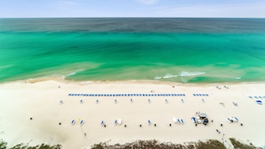 An aerial view of a beach with rows of sun loungers and umbrellas, a clear sky, and the ocean in the background.