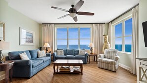 A living room with a blue sofa, a striped armchair, a wooden coffee table, and a ceiling fan. There are two windows with curtains, a lamp, and a vase on a side table.