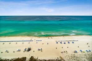 An aerial view of a beach with rows of blue and white sun loungers, a few beach umbrellas, and a small building likely for beach activities. - Beach Service Included | Gulf-Front 15th Floor Condo | Stunning Sunset Views (Panama City Beach)