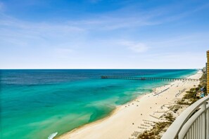 En strand med en brygga som sträcker sig ut i havet, en sandstrand och en byggnad med balkonger som vetter mot landskapet.