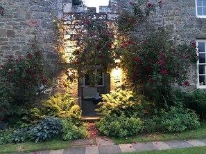 Enter a lush garden, surrounded by a variety of plants and flowers, leading to a historic building with a rustic charm.