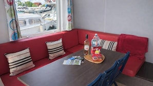 A dining area with a table set for two, featuring a bottle of water, a bottle of wine, and a packet of snacks. The room has red seating and a window with a view of boats.