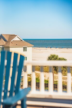 A beach view from a balcony with a chair in the foreground, a building with a sloped roof, and the ocean in the background.