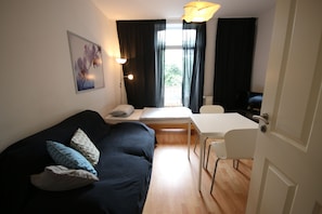 A compact living space with a dark-colored sofa, a small dining table, and a white chair. A painting is visible on the wall, and there is a window with curtains. - a-domo Apartments Essen - Serviced Apartments & Flats - short or longstay - single or grouptravel (Essen)