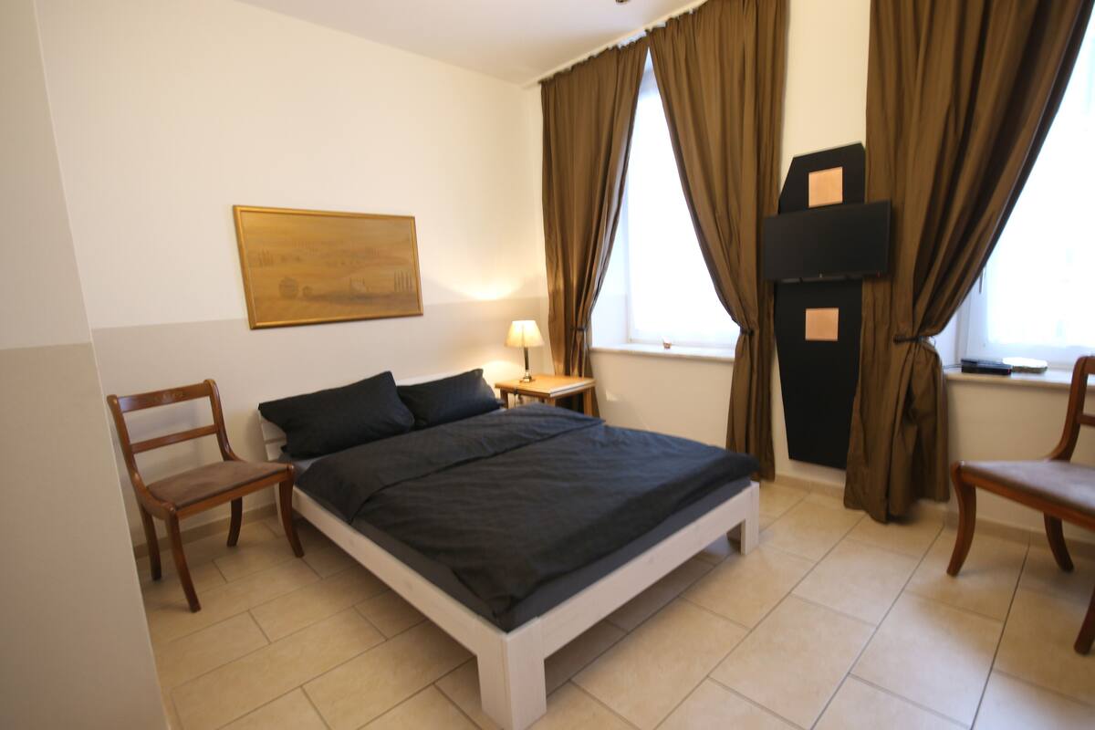 A bedroom with a bed, two chairs, a nightstand with a lamp, a TV, and a painting on the wall.
