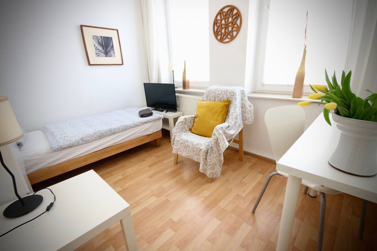 A small, tidy room with a single bed, a chair, a desk, and a potted plant.