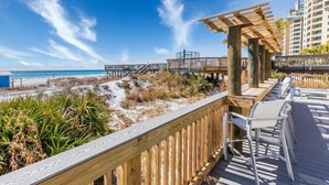 Une terrasse en bois offrant une vue sur la plage et l’océan, un espace salon couvert et un bâtiment en arrière-plan.