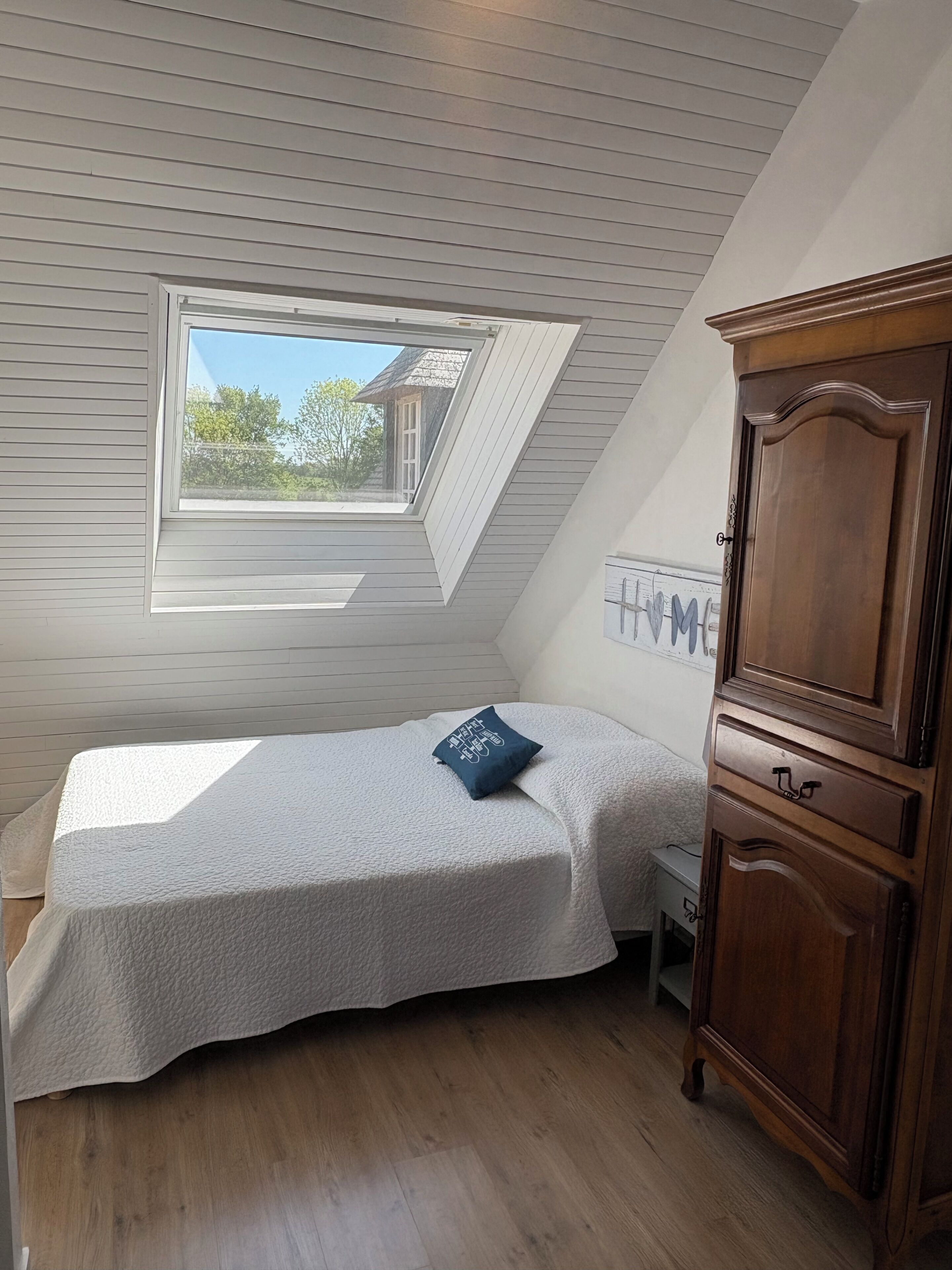 A bedroom with a sloped ceiling, a skylight, a bed with a pillow, a wooden wardrobe, and a wall-mounted TV.