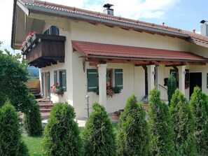 Une maison à deux étages, avec un toit recouvert de tuiles rouges, un balcon doté de balustrades en bois et une véranda couverte avec des volets verts.