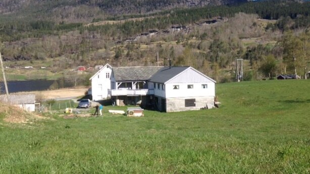 Ett vitt hus i två våningar med en balkong, beläget på en gräsklädd äng med ett berg i bakgrunden.