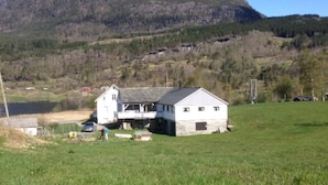 Ein weißes Gebäude mit Balkon, eine kleine Gartenhütte und ein Auto auf einer Wiese, im Hintergrund ein Berg.