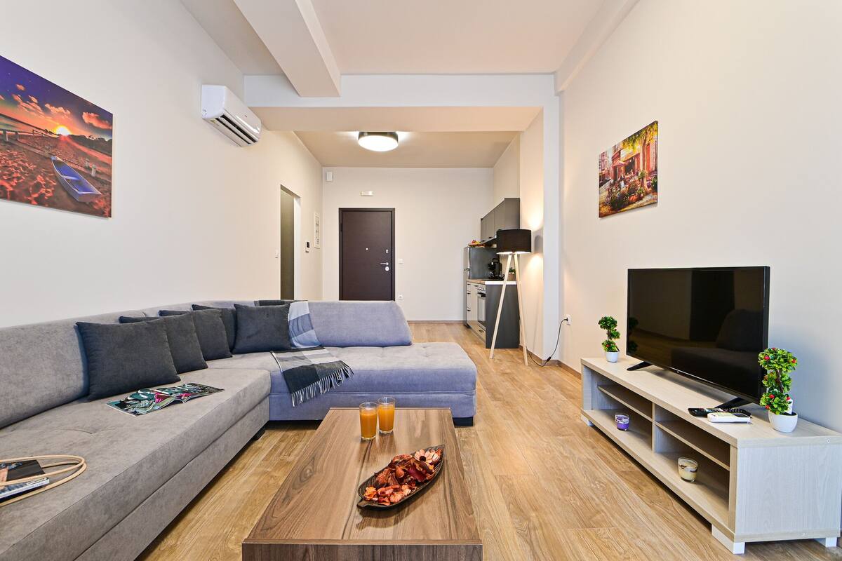 Una sala de estar moderna con un sofá gris, una mesa de centro de madera con una bandeja de aperitivos, un televisor de pantalla plana sobre un mueble bajo y una pequeña planta en maceta.