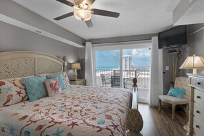 A bedroom with a bed, a ceiling fan, a TV, a balcony with a view of the ocean and a building.