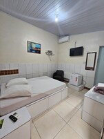 Quarto de hotel com cama, poltrona, televisão, ar condicionado e mesa pequena com controle remoto.