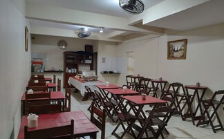 Uma sala de jantar com várias mesas e cadeiras, uma cozinha ao fundo e um relógio na parede.