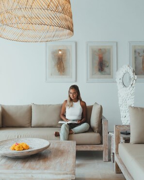 En person sitter i en soffa och läser en bok i ett rum med en vävd lampa, inramade konstverk på väggen och en dekorativ vit väggdekoration.