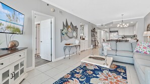 A spacious living room with a large TV, a sofa, and a dining area with a chandelier. - The Enclave 203 | Gulf Front Gem | Incredible Views from Private Balcony | My Beach Getaways (Orange Beach)
