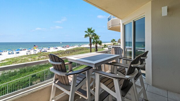 A balcony with a table and chairs, overlooking a beach with palm trees and a clear blue sky. - The Enclave 203 | Gulf Front Gem | Incredible Views from Private Balcony | My Beach Getaways (Orange Beach)