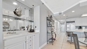 A modern kitchen with white cabinetry, a large island with bar stools, a decorative metal shelf, and a wall-mounted mirror. - The Enclave 203 | Gulf Front Gem | Incredible Views from Private Balcony | My Beach Getaways (Orange Beach)