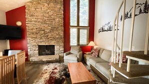 A cozy living room with a stone fireplace, a red accent wall, a comfortable sofa, a wooden coffee table, and a staircase with a railing.