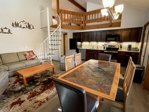 Un vaste espace de vie doté d’un haut plafond, d’un escalier en bois, d’une table à manger avec des chaises, d’un canapé et d’une cuisine à l’arrière-plan.