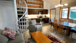 A modern living room with a spiral staircase, a dining area with a wooden table and chairs, and a kitchen with dark cabinets and stainless steel appliances.