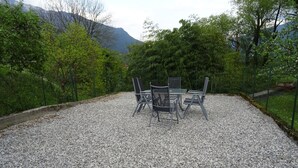 An outdoor seating area with a gravel surface, surrounded by greenery and a mountain backdrop. - Hiša Kramer - Apartment Sedlo (Kobarid)