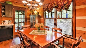 A wooden cabin interior with a dining table, chairs, a chandelier, and a window with curtains.