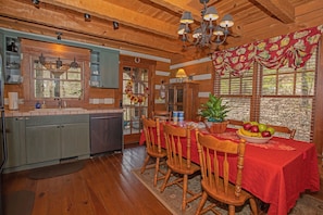 Interior de uma cabana de madeira com cozinha, área de jantar e uma grande janela com persianas.