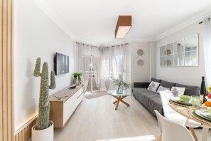 Een moderne woonkamer met een bank, een eettafel met stoelen, een cactus en een aan de muur bevestigde televisie.