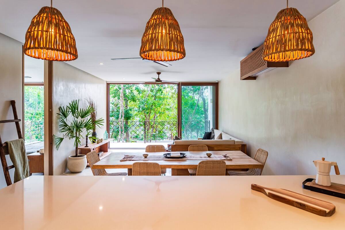 Un espace repas avec une grande table, trois lampes suspendues en osier et une vue sur la verdure grâce à de grandes fenêtres.
