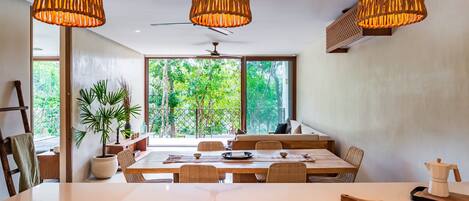Un comedor con una mesa grande, tres lámparas de mimbre colgantes y vistas a vegetación a través de ventanas amplias.
