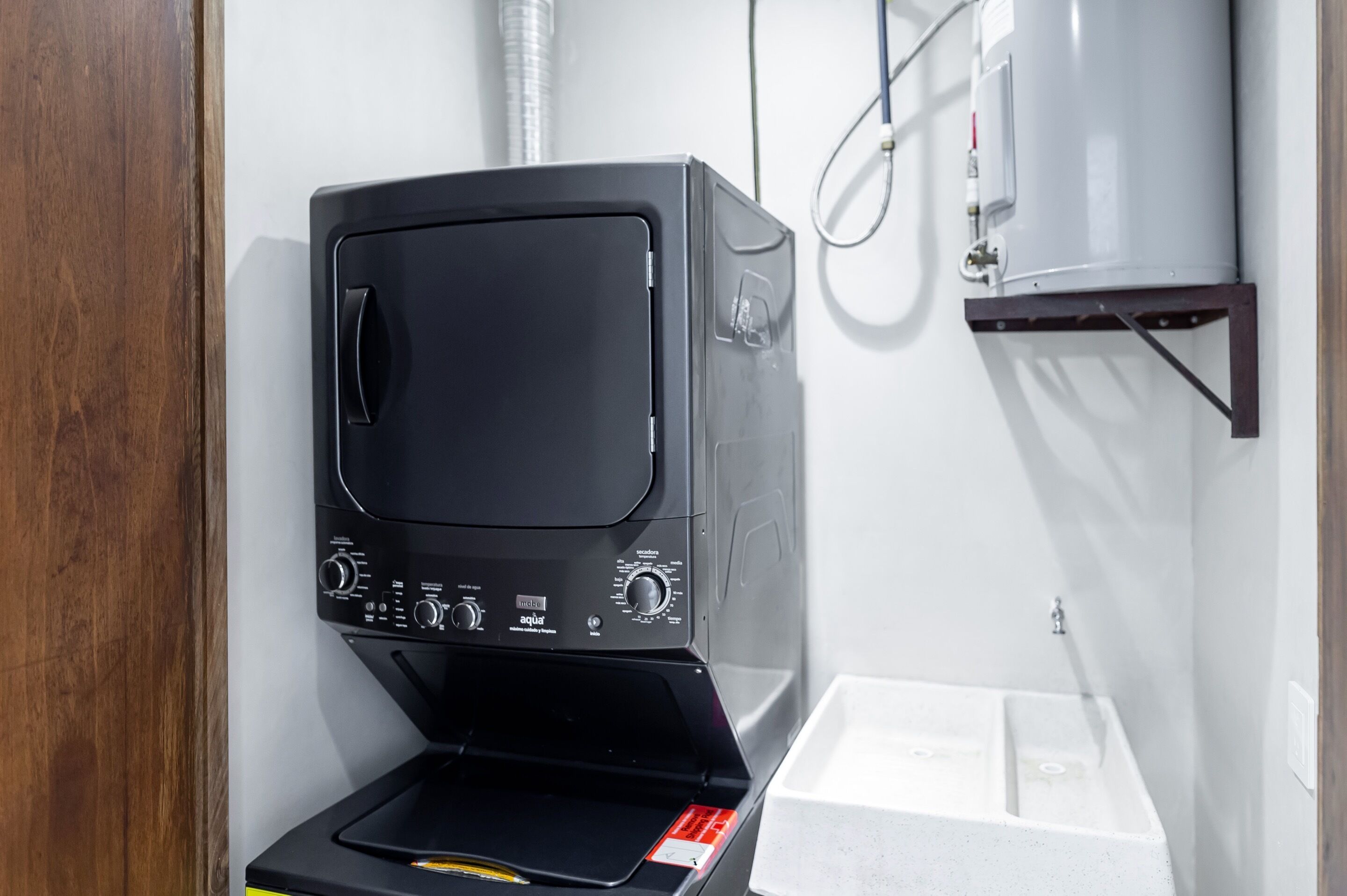 A black washing machine in a small room with a white sink and a water heater mounted on the wall.