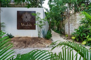 Une enseigne pour Wahh Tulum, avec un motif géométrique, entourée d’une végétation luxuriante et d’un mur de pierre.