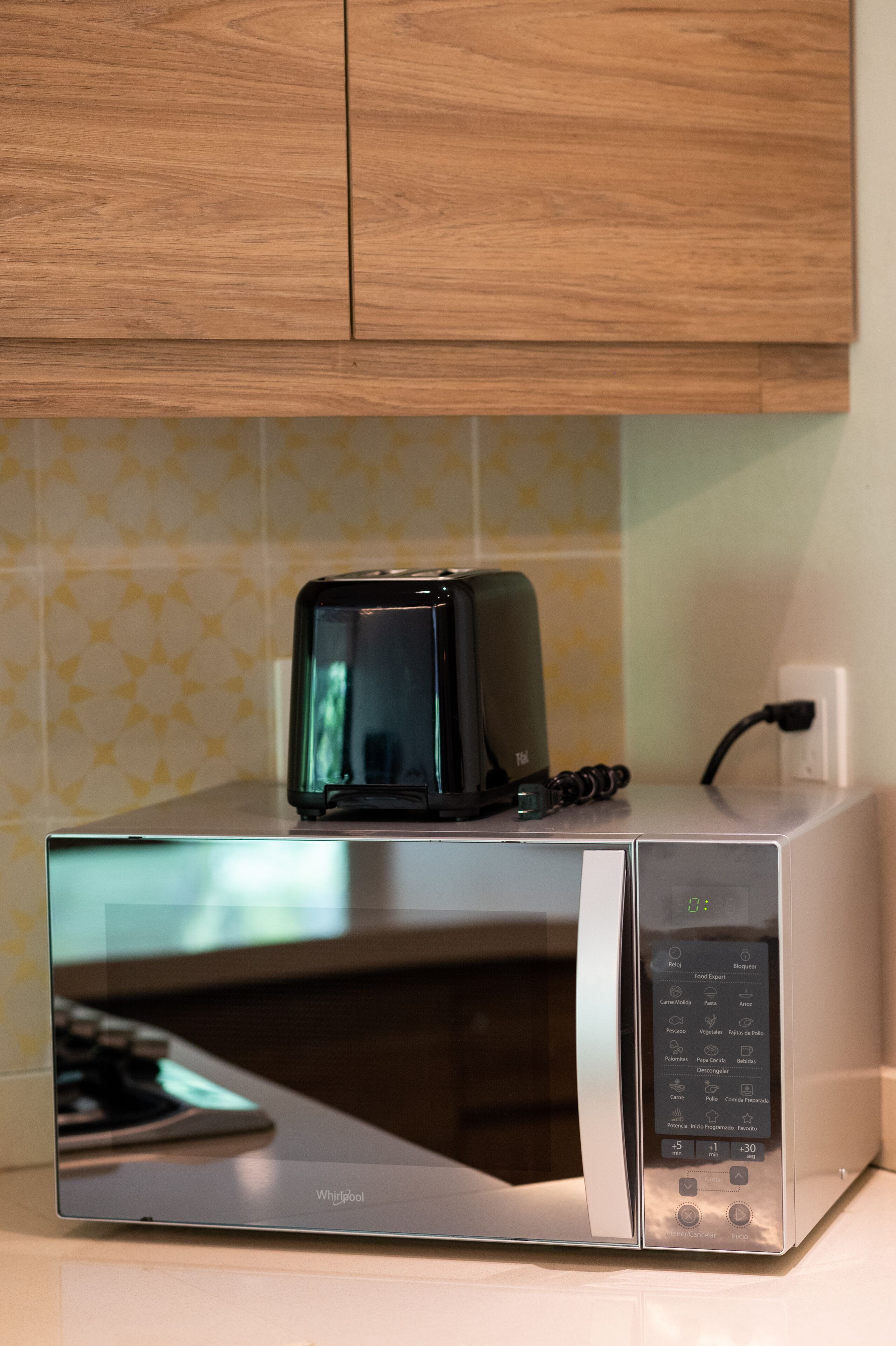 A compact microwave oven with a digital control panel and a toaster on top, placed on a countertop with a patterned backsplash.