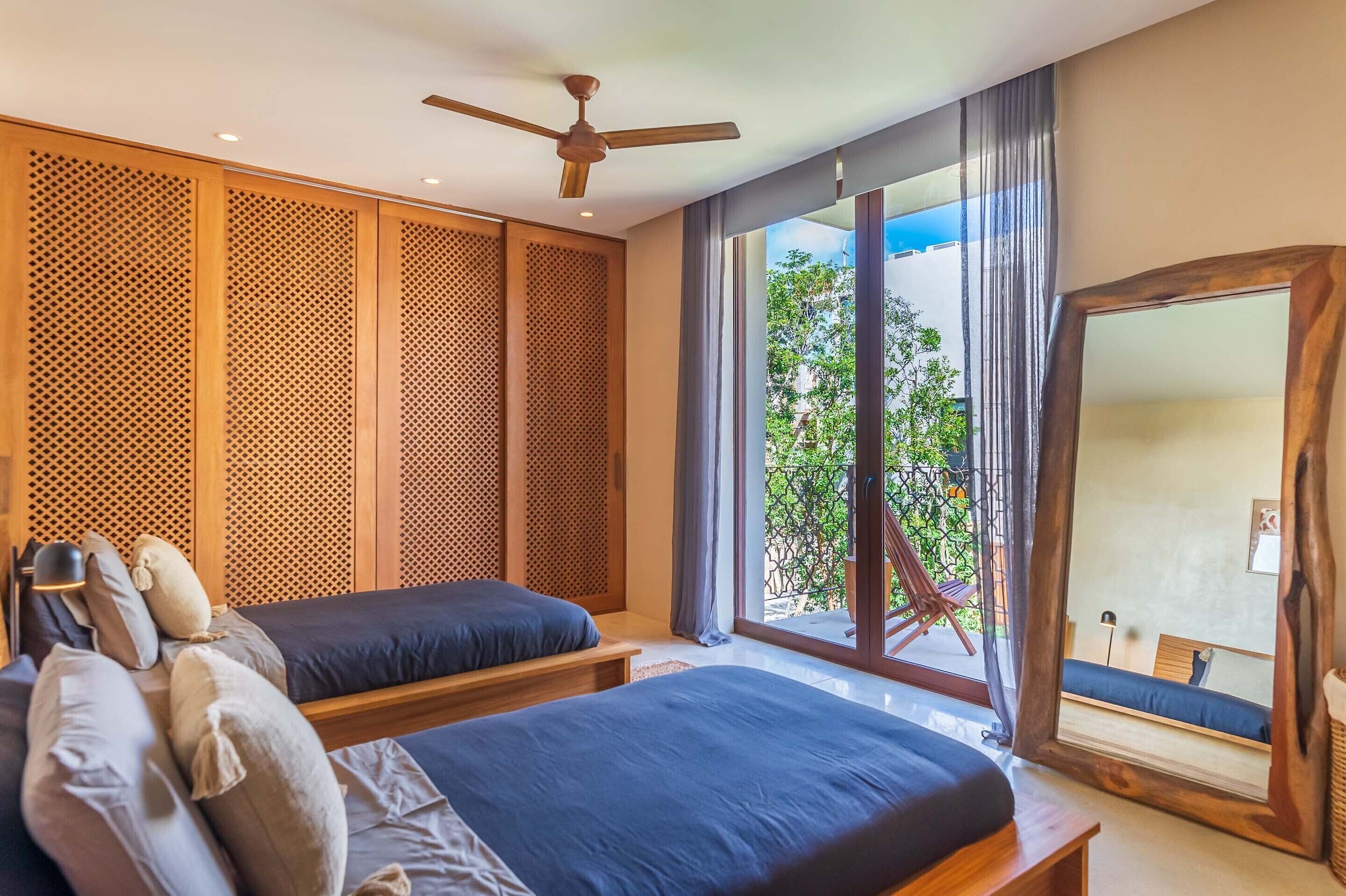 A bedroom with two beds, a ceiling fan, a large mirror, and a view of greenery outside.
