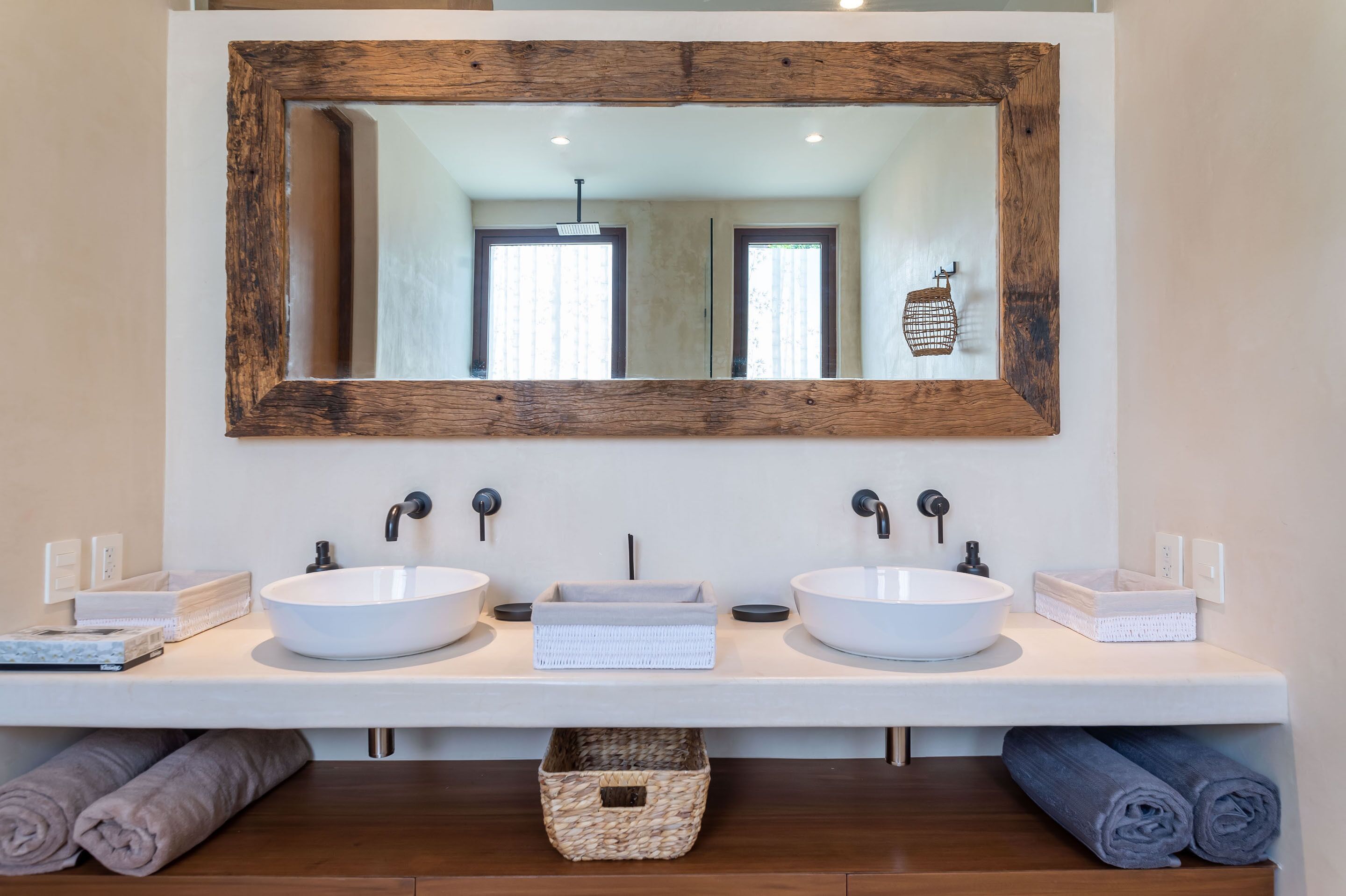A bathroom with two white sinks, a large mirror with a rustic frame, and towels neatly rolled up.