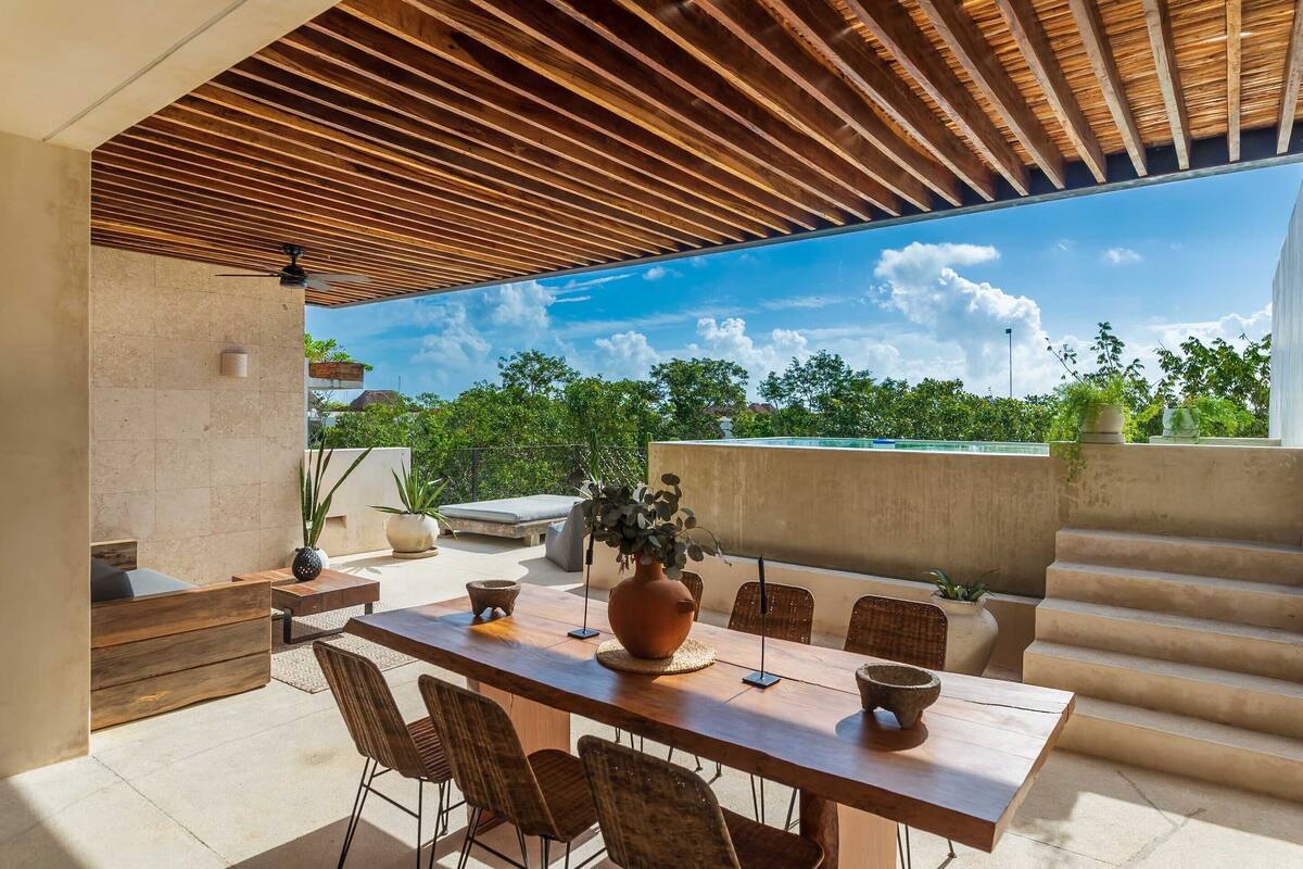Une terrasse extérieure avec une table et des chaises en bois, un grand vase et une vue sur la verdure et une piscine.