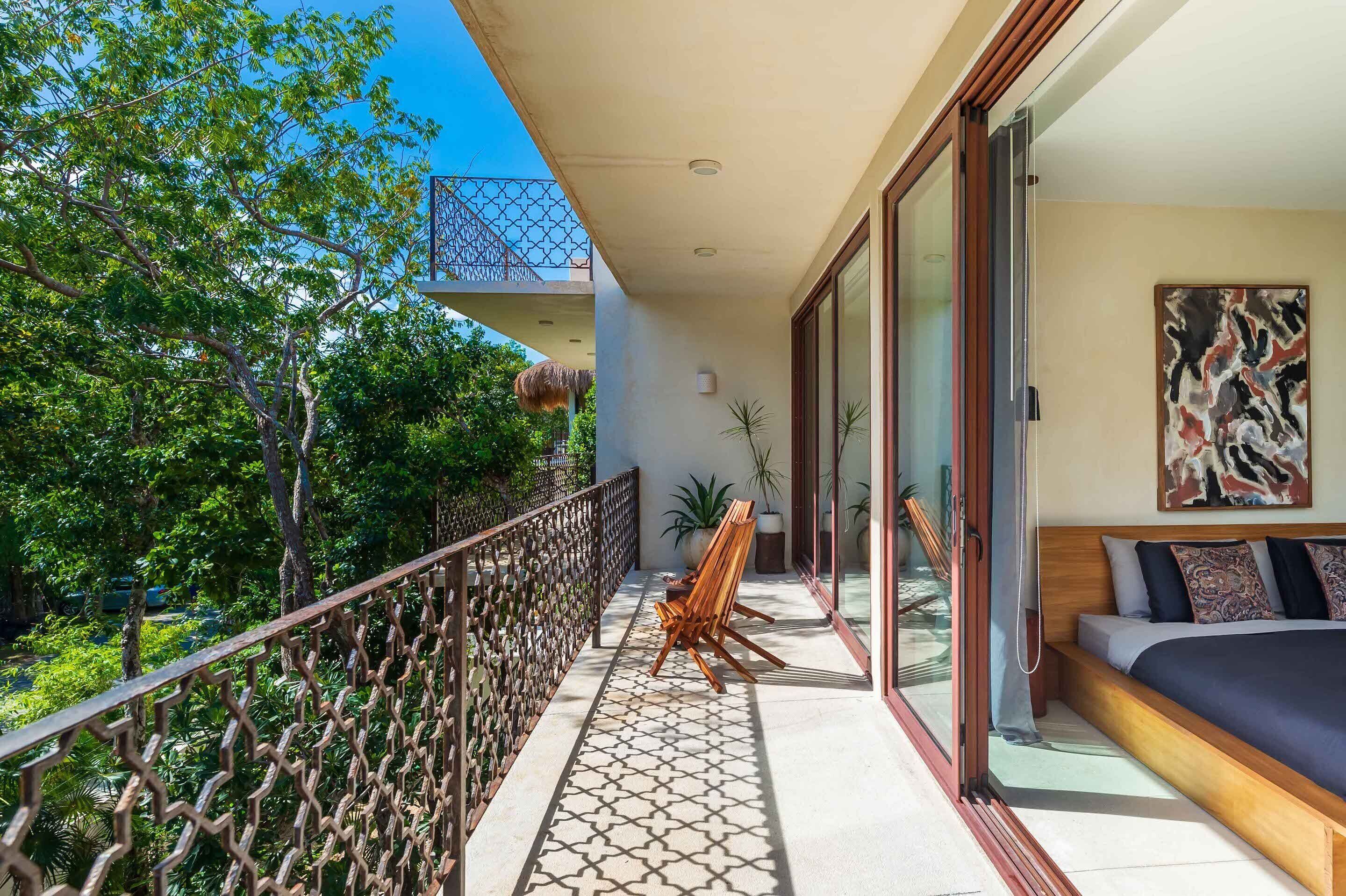 A balcony with a wooden chair, a bed with a patterned bedspread, and a view of trees and a thatched roof structure.