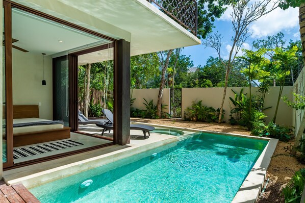 Un espace piscine extérieur moderne doté d’un salon clos par des baies vitrées, d’une terrasse en bois et offrant une vue sur une végétation luxuriante.
