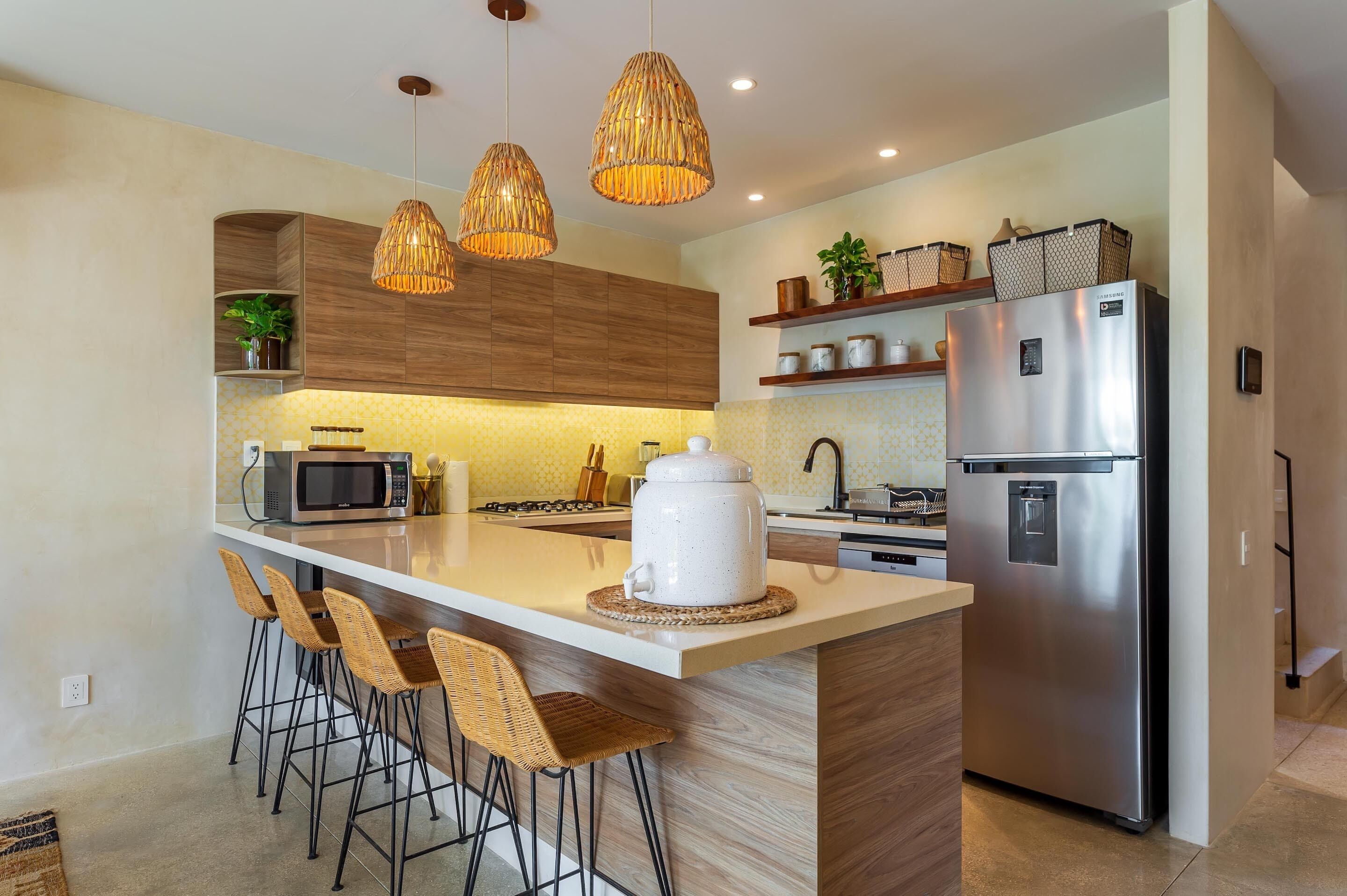 A modern kitchen with a central island, bar stools, a stainless steel refrigerator, and wicker pendant lights.