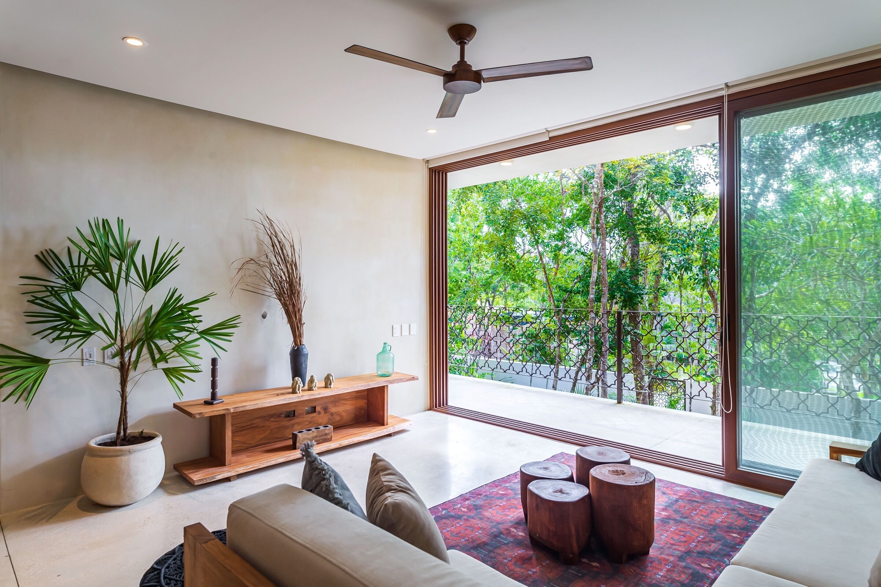Un salon moderne doté d’une grande porte-fenêtre coulissante donnant sur un balcon, d’un ventilateur de plafond et d’une console en bois agrémentée d’objets décoratifs.