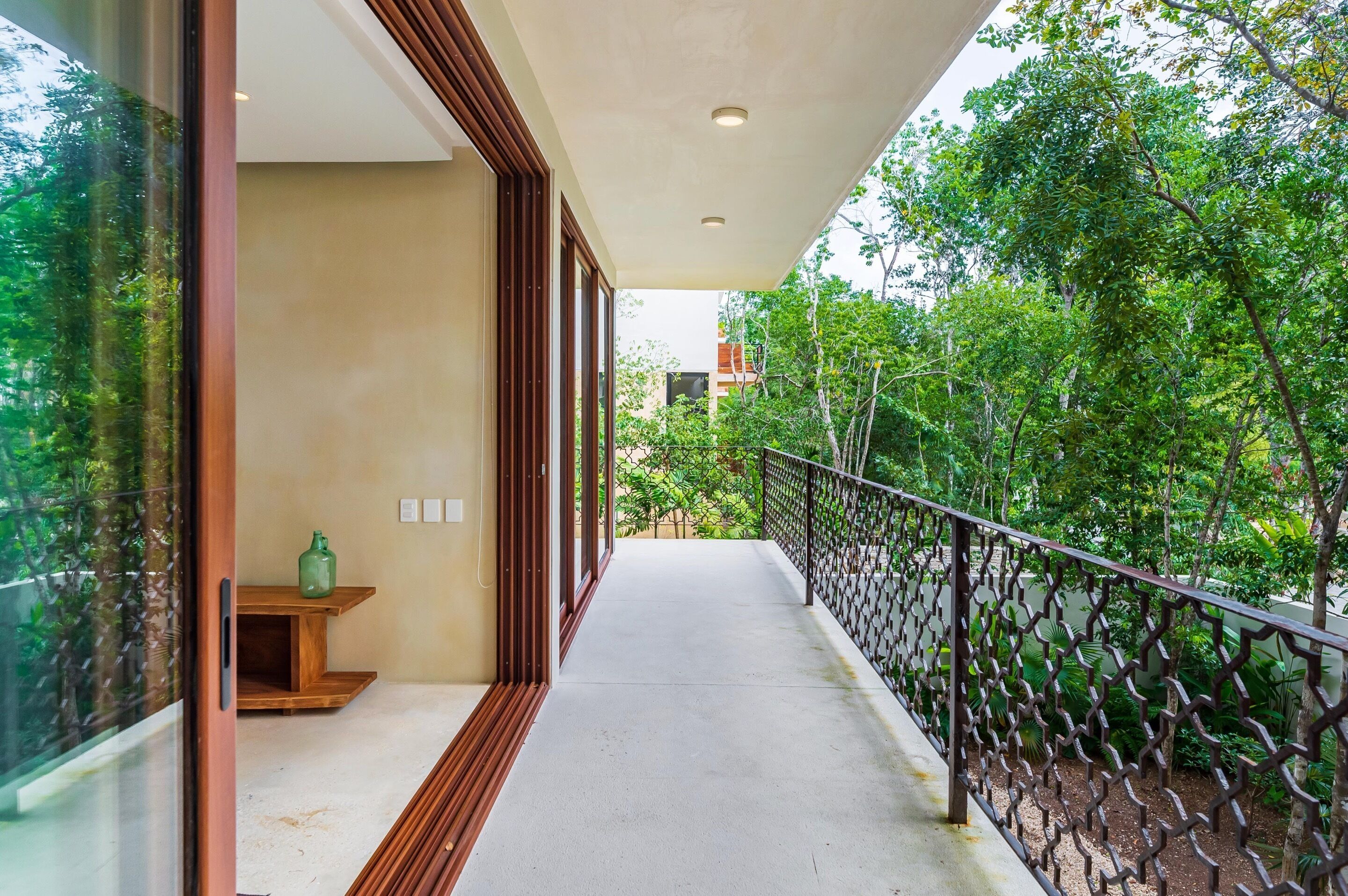 A balcony with a view of lush greenery, a wooden bench, and a glass door leading to a room.