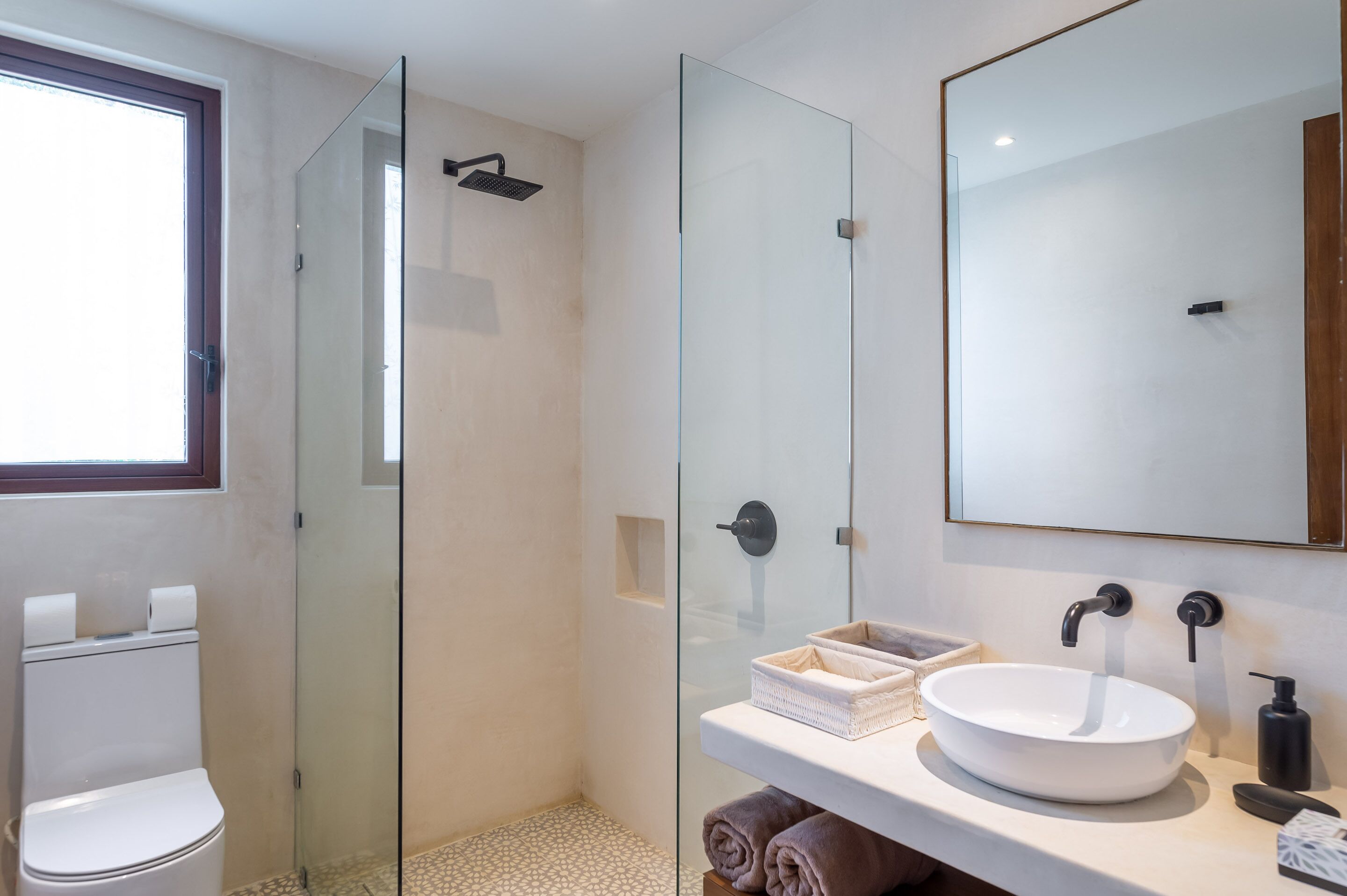 A modern bathroom with a glass-enclosed shower, a white sink with a large mirror, and a toilet with a window above it.