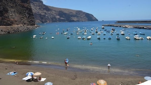Ein Strand mit zahlreichen Booten, die im Wasser ankern, einer felsigen Klippe an einer Seite und einem Steg, der ins Meer ragt.