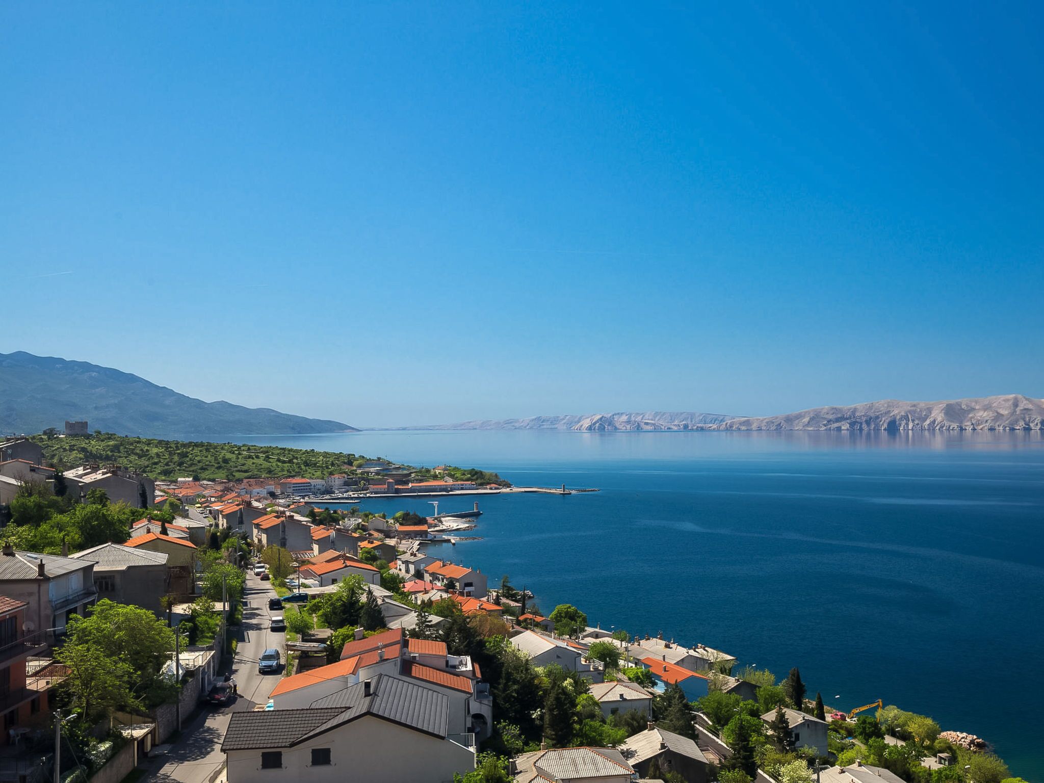 A coastal town with buildings along the waterfront, a clear blue sky, and mountains in the background.