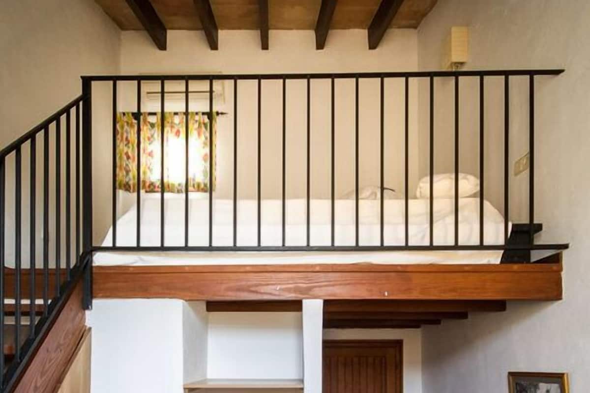 A loft bedroom with a wooden ceiling, a black railing, and a view of a window with curtains.