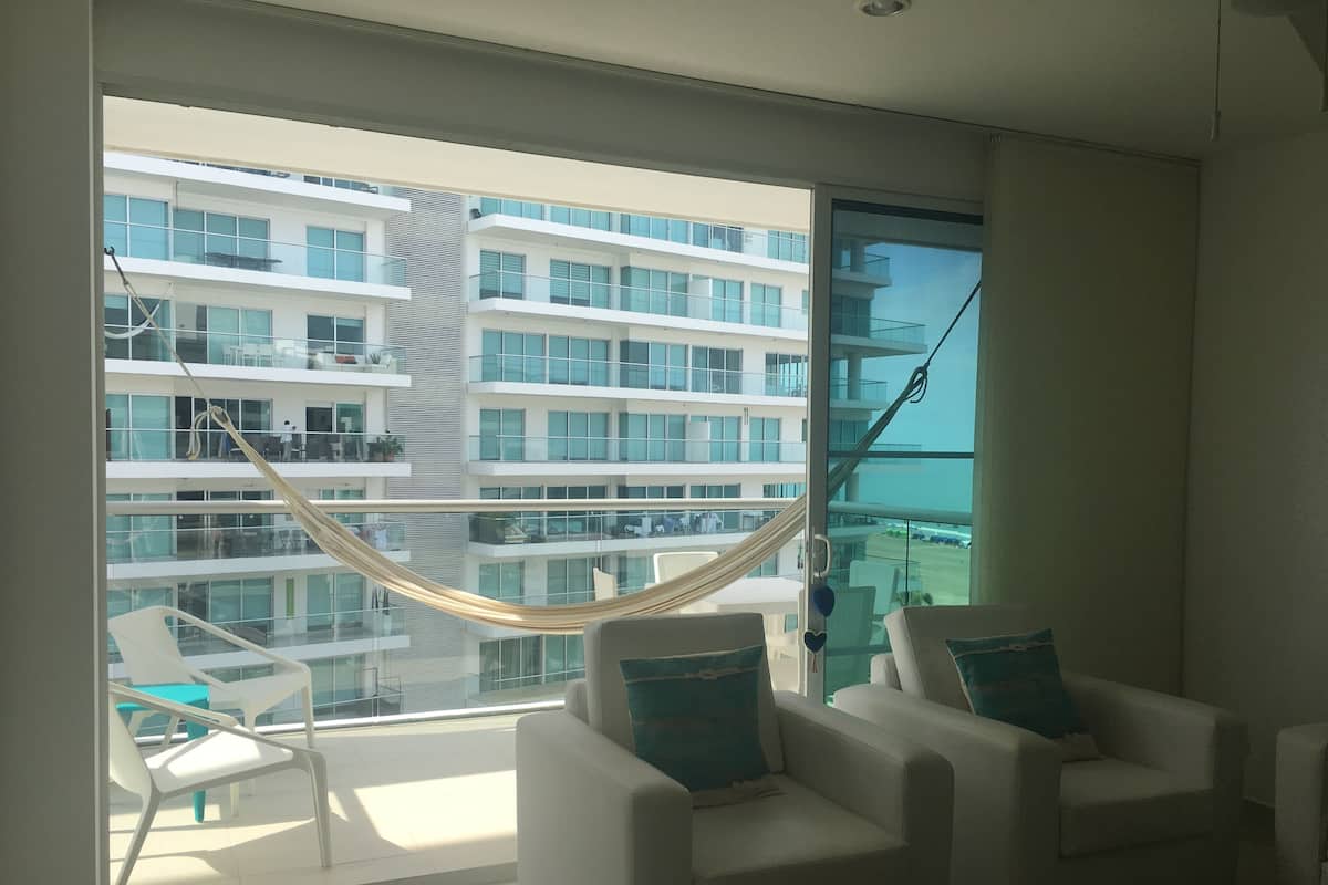 A modern hotel room with a large balcony, white furniture, and a view of a high-rise building.